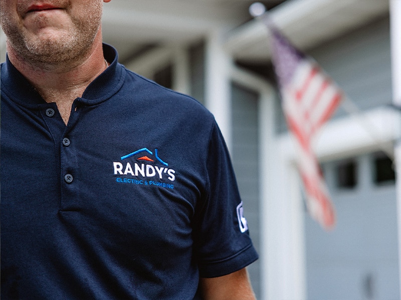 Man in Randy's Electric & Plumbing shirt near American flag and house.