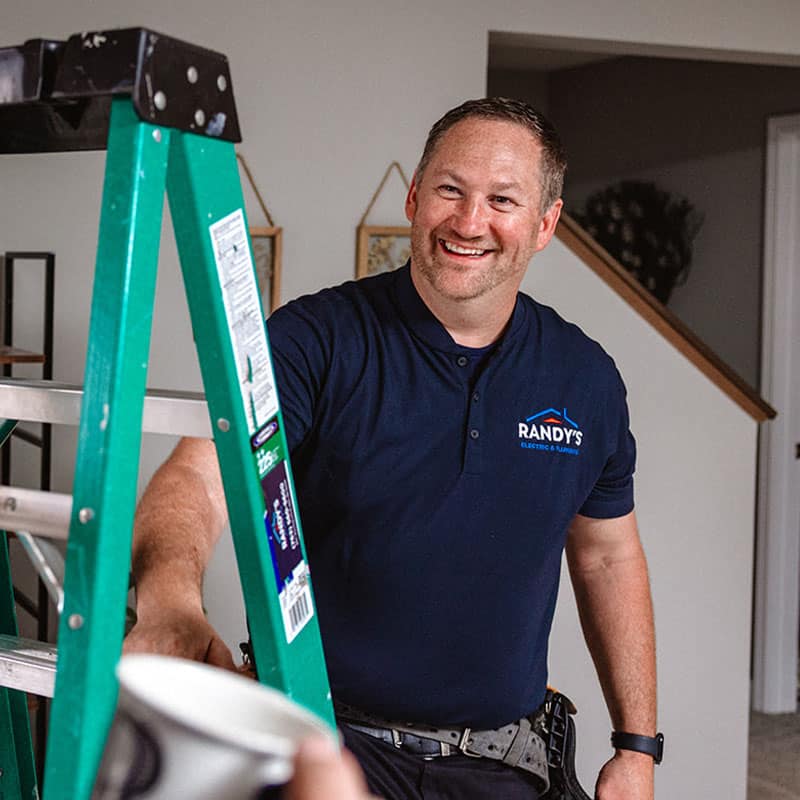 Man in a Randy's Electric shirt smiling by a green ladder indoors.
