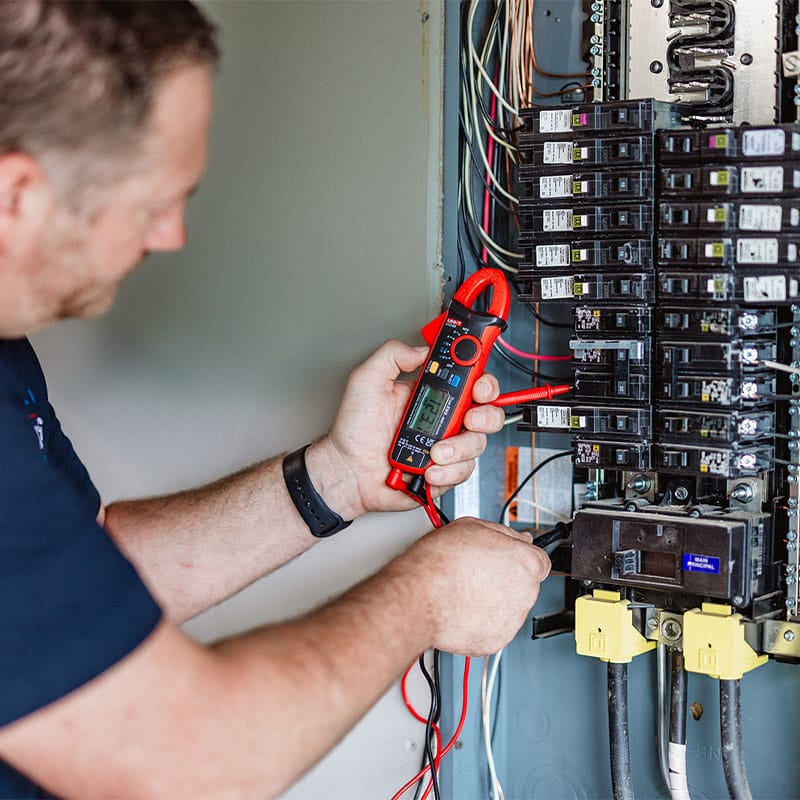 Electrician using a digital multimeter to test an electrical panel for voltage readings.