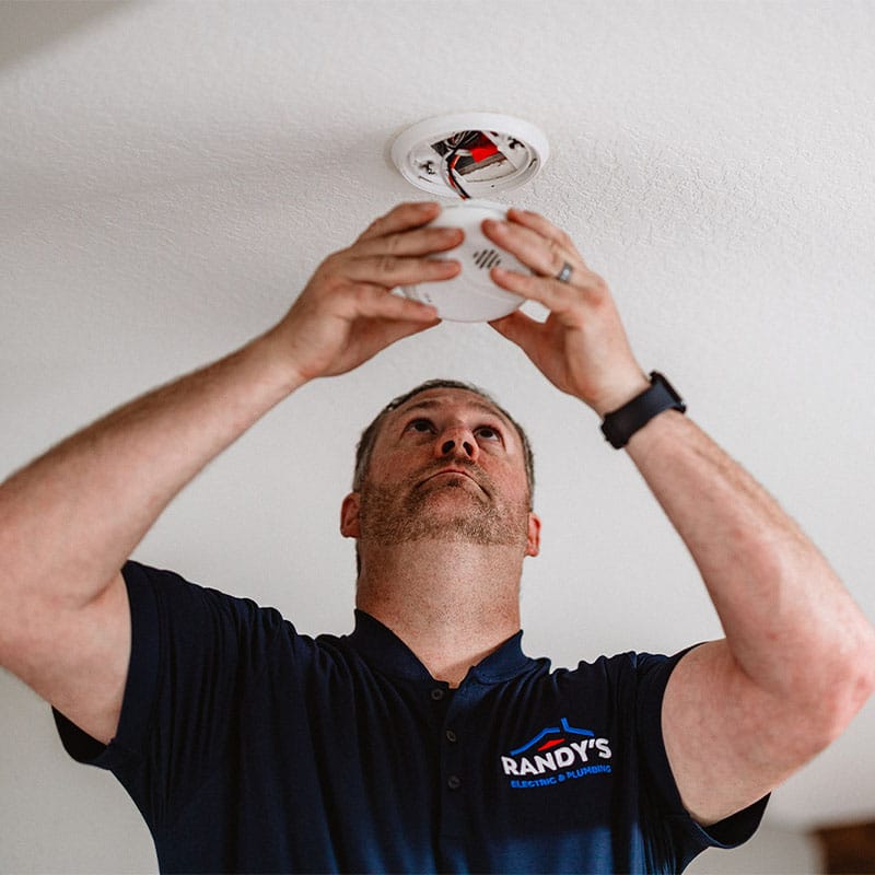 A man from Randy's Electric & Plumbing installs a smoke detector on the ceiling.