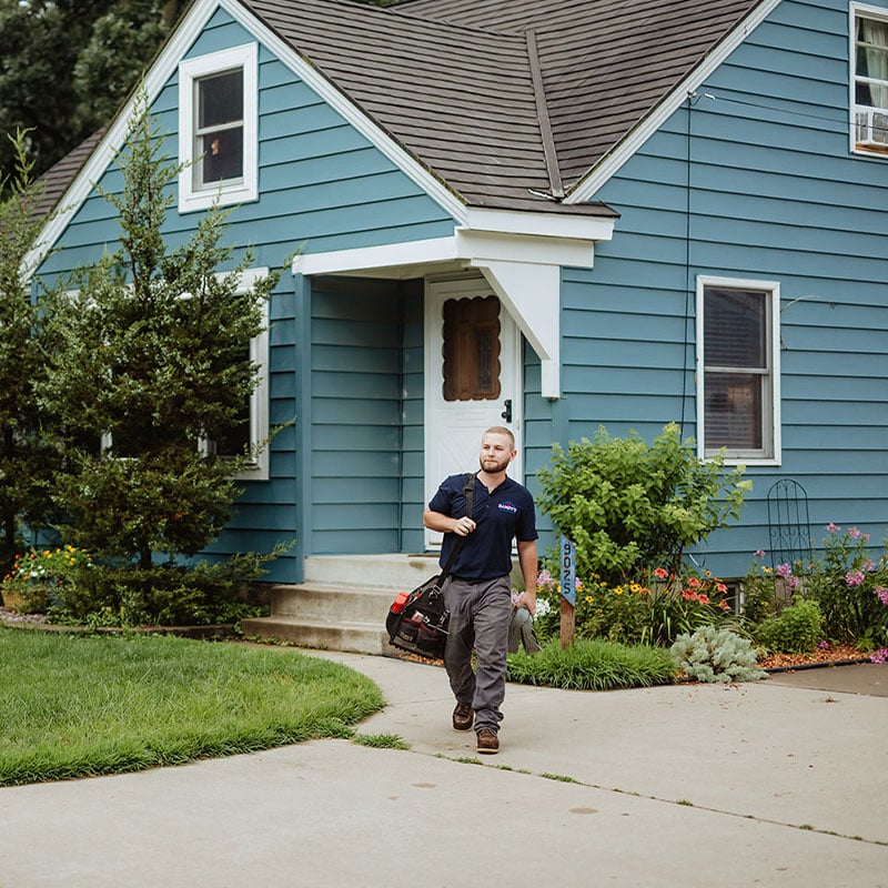 A man carrying a tool bag walks outside a blue house.