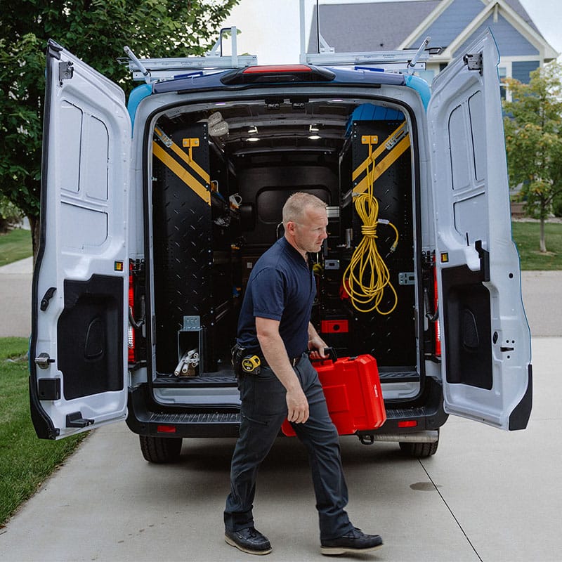 A worker carrying a toolbox from the back of a service van, driveway and houses in the background.