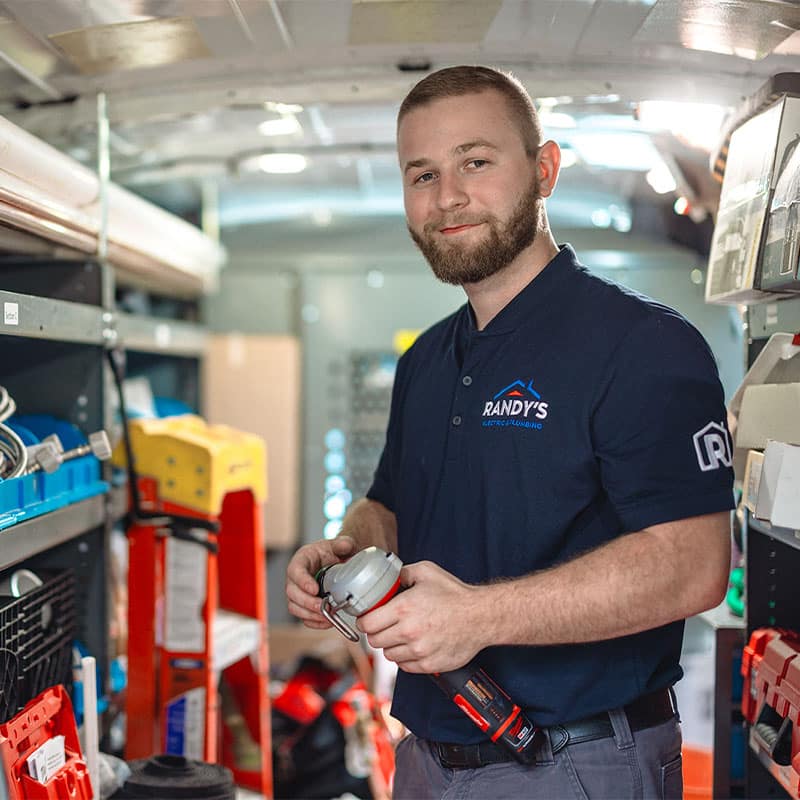 Man in a Randys Electric & Plumbing shirt inside a work van, holding a tool, surrounded by equipment.