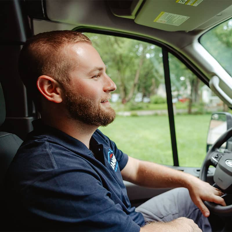 A man driving a van, smiling, in a navy blue uniform with logo, green trees visible through the window.