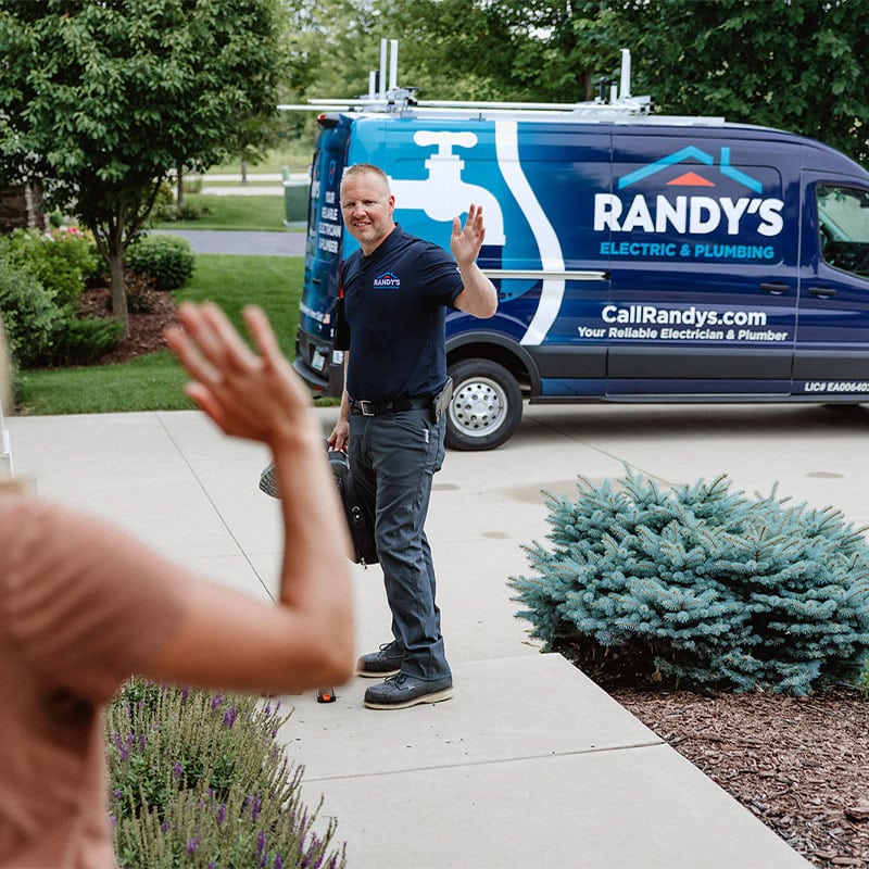 Randy waving outside home with electrician and plumbing van in driveway.