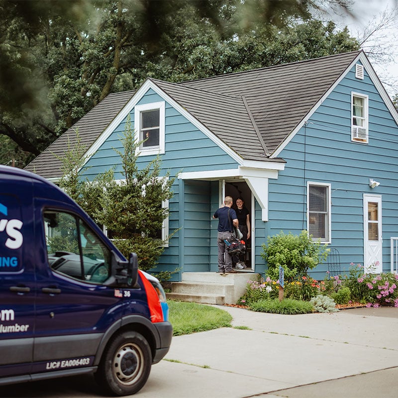 Plumber at a blue house entrance, parked van displaying company branding, amidst greenery and flowers.
