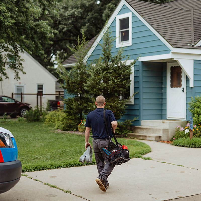 A technician walking towards a blue house carrying tools and equipment.