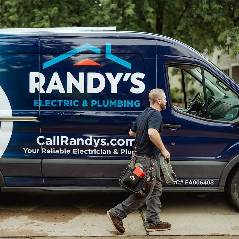 A technician walking past a Randy's Electric & Plumbing van, carrying tools and equipment.
