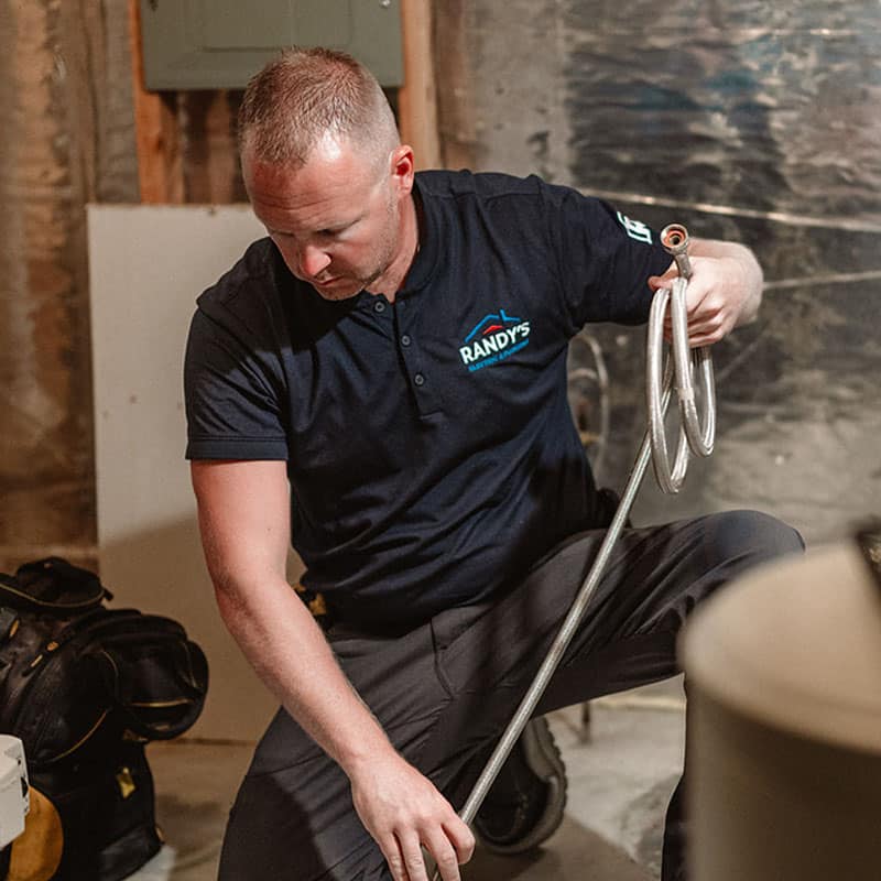 Randy kneeling in a basement holding coiled metal pipes, wearing a dark shirt with company logo.