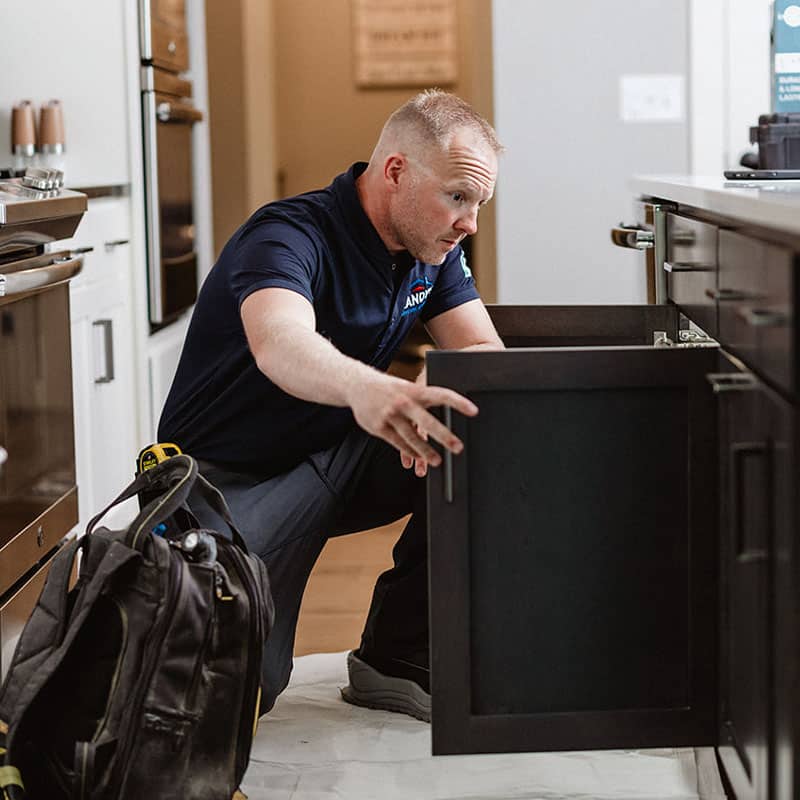 A plumber examining a kitchen sink cabinet, tools in a bag nearby.