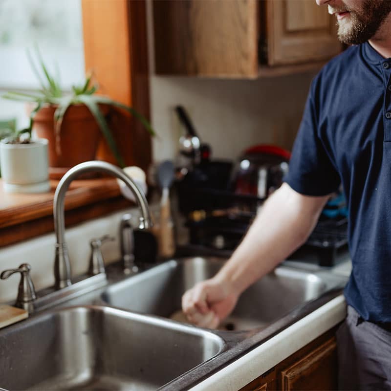 A man in a navy shirt washing dishes at a kitchen sink with plants on the windowsill.