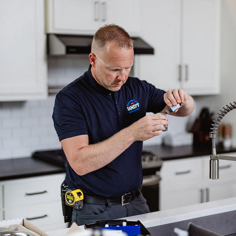 A man in a kitchen assembling a small object, wearing a shirt with RANDY'S logo.