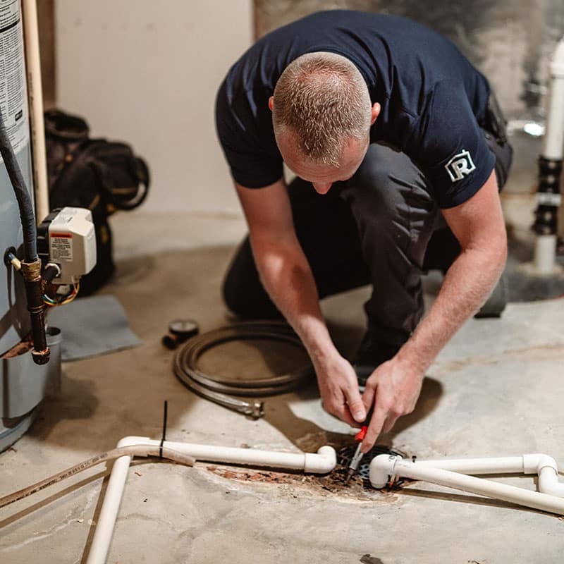 Man repairing pipes in a basement, using pliers and wearing a navy blue shirt.