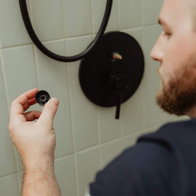 Person inspecting a piece of plumbing fixture beside a shower handle in a bathroom.