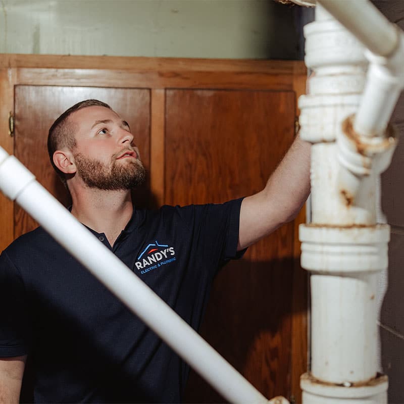 Man inspecting plumbing pipes in a basement, wearing a Randy's shirt.