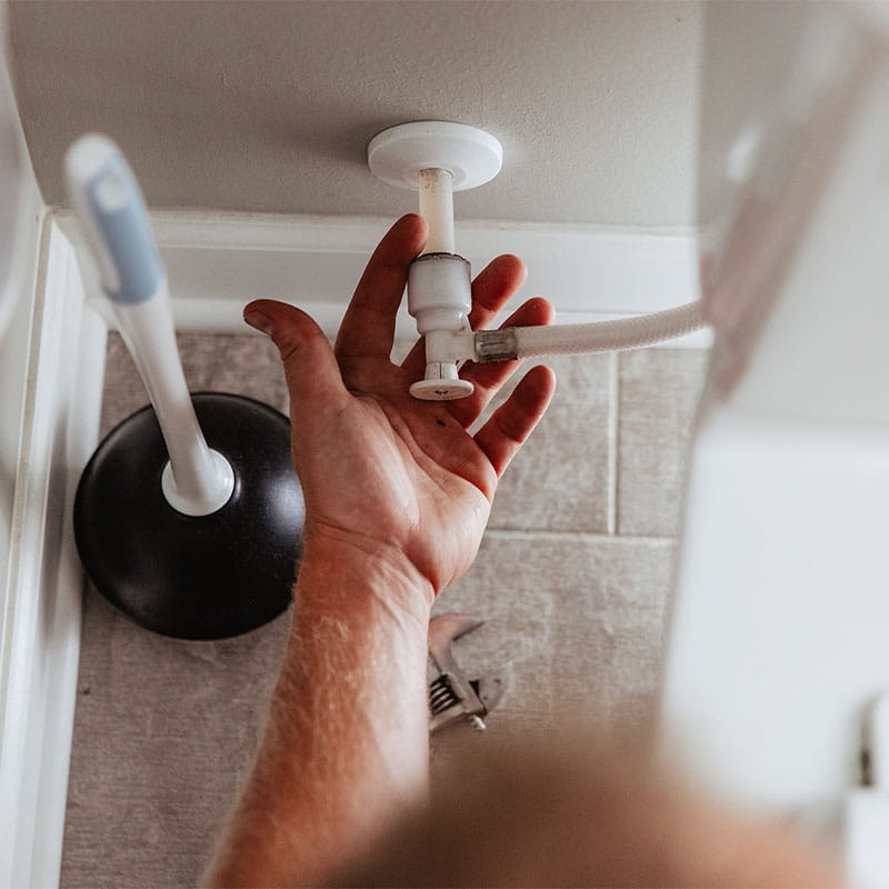 Hand fixing a pipe beneath a sink with a wrench, plunger nearby.