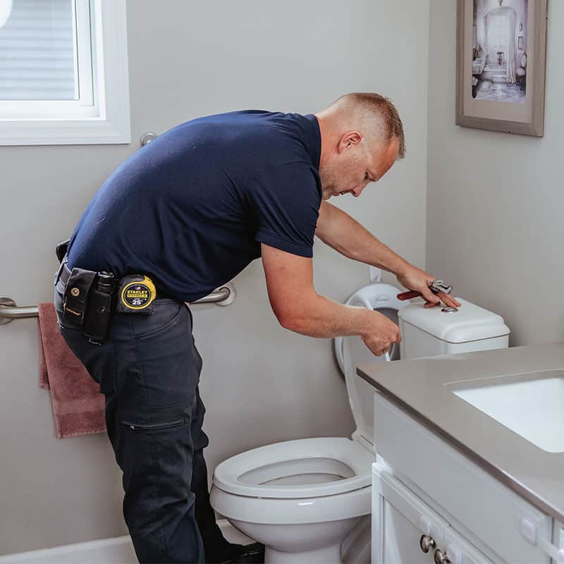 A plumber in a blue shirt fixing a toilet in a bathroom setting.