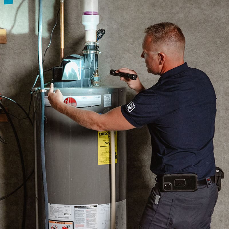 A worker inspects a water heater with a flashlight in a utility room.