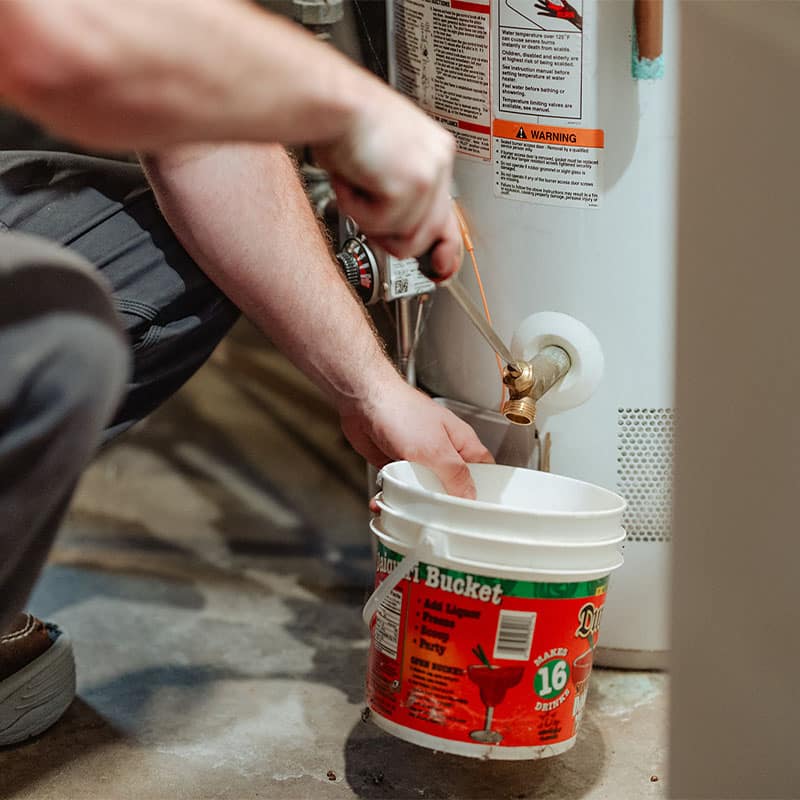 Person draining a water heater into a white bucket in a basement.