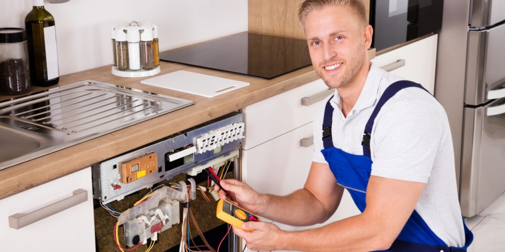 Electrician repairing kitchen appliance, kneeling and smiling, wearing blue overalls.