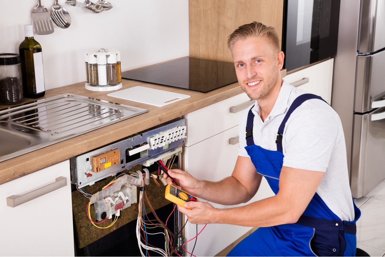Electrician repairing kitchen appliance, kneeling and smiling, wearing blue overalls.