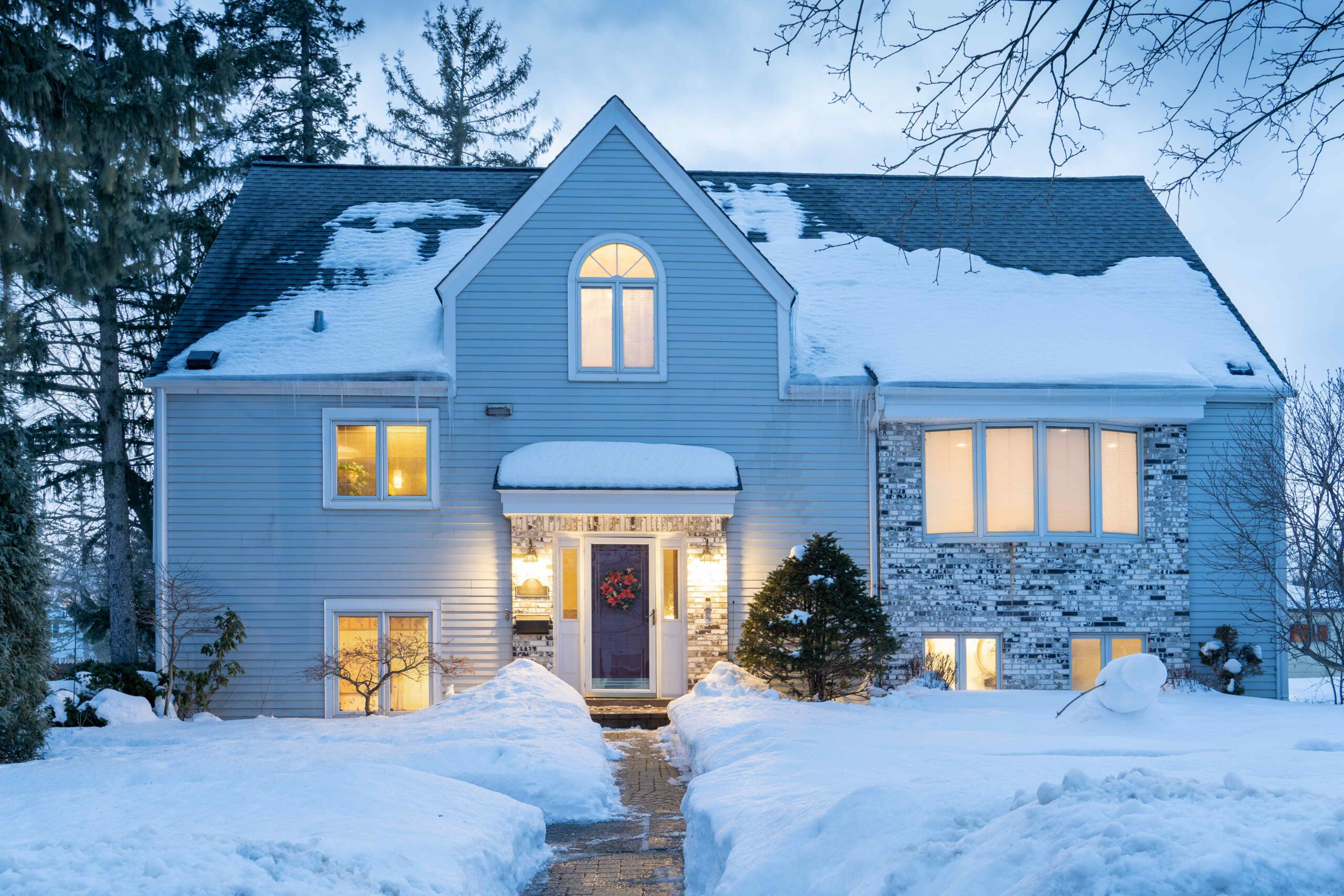 Snow-covered house with lit windows at dusk, surrounded by trees and a path through the snow.