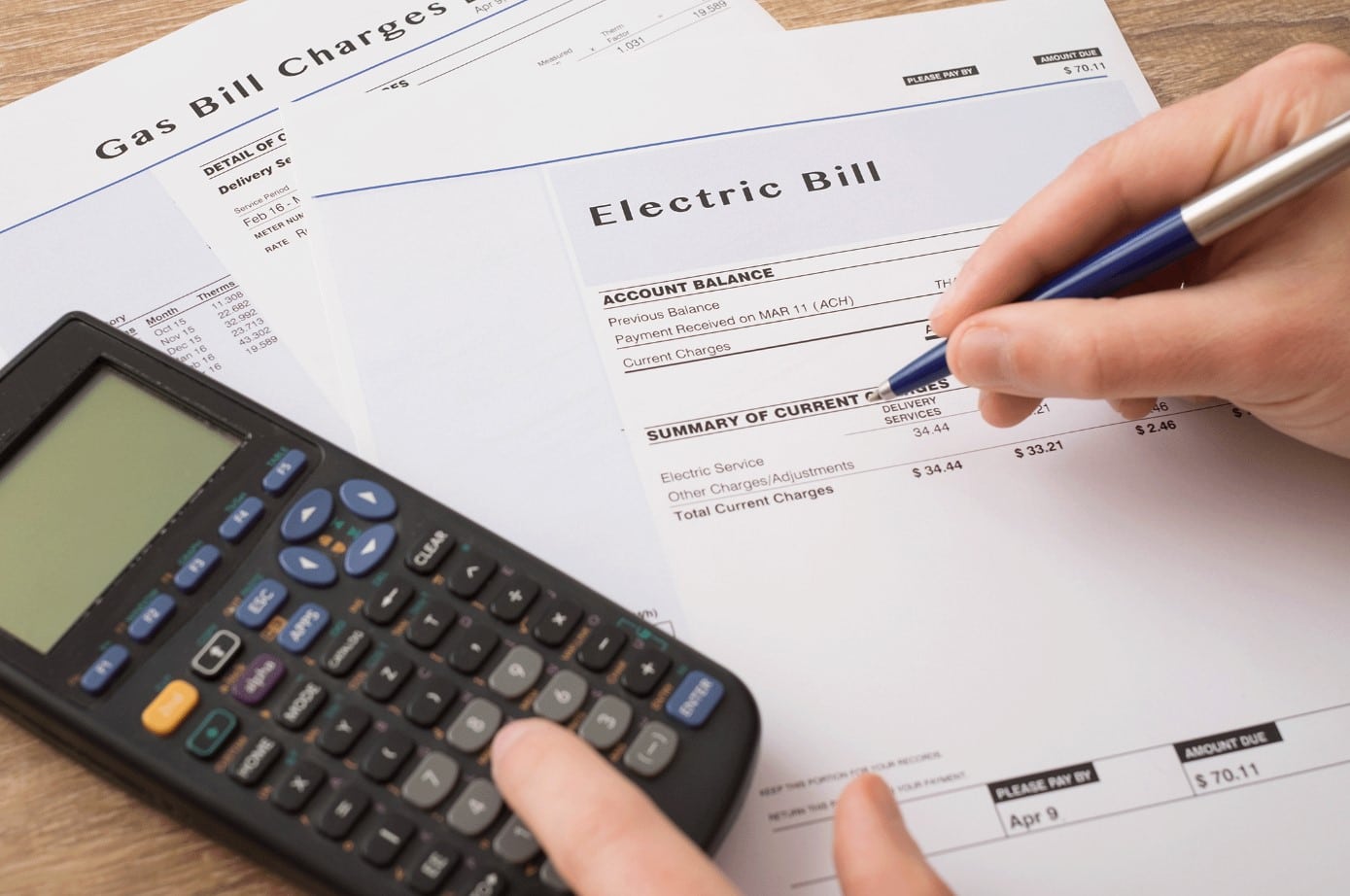 Hands with pen and calculator reviewing electric and gas bills on a wooden desk.