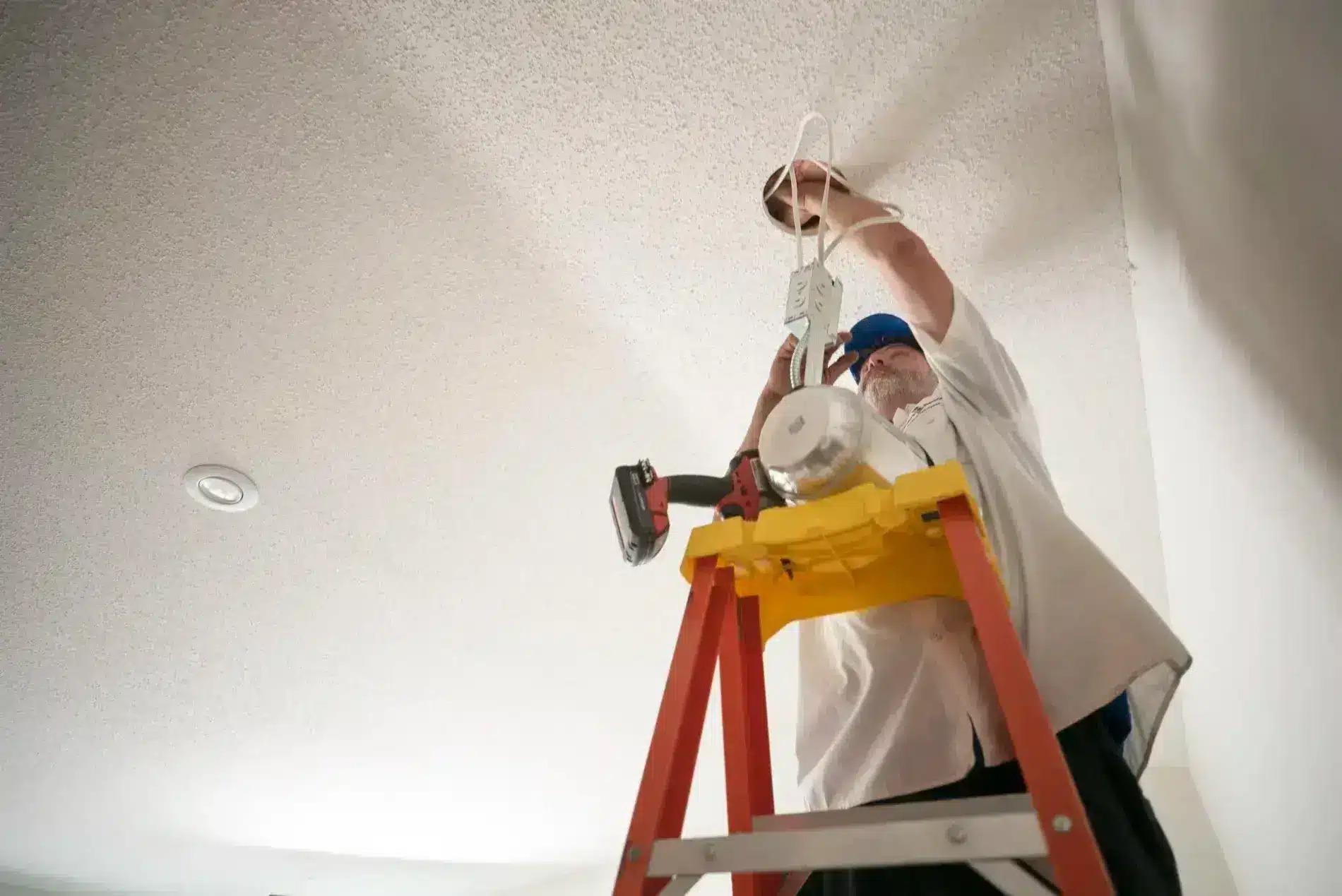 Electrician on ladder installing ceiling lighting fixture.
