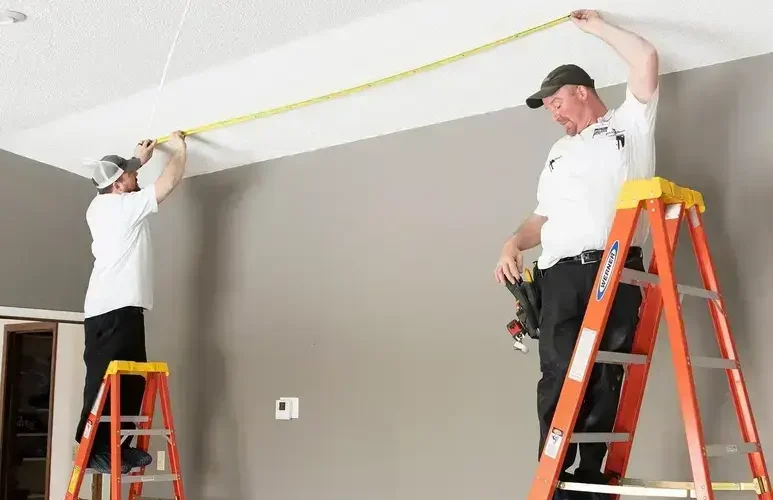 Two workers on ladders measuring a ceiling with a tape measure in a room.