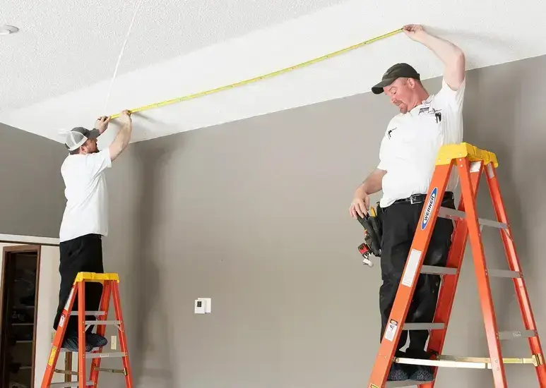 Two workers on ladders measuring a ceiling with a tape measure in a room.