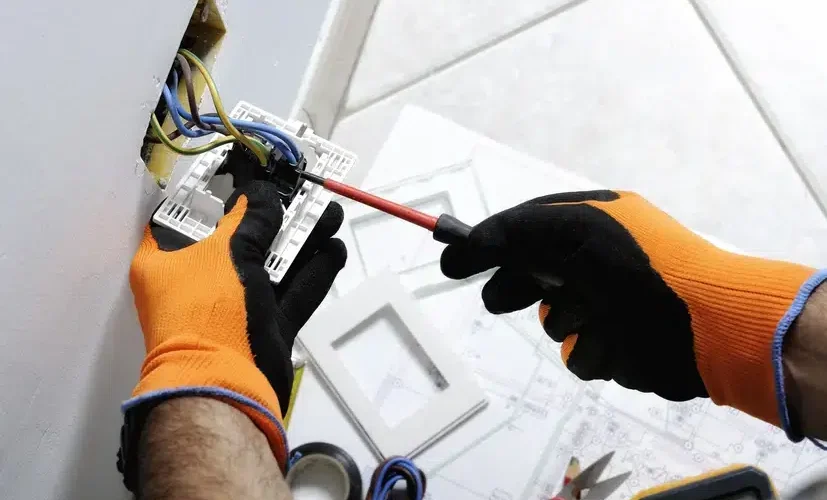 Electrician in orange gloves using a screwdriver to fix a wall socket with exposed wires.