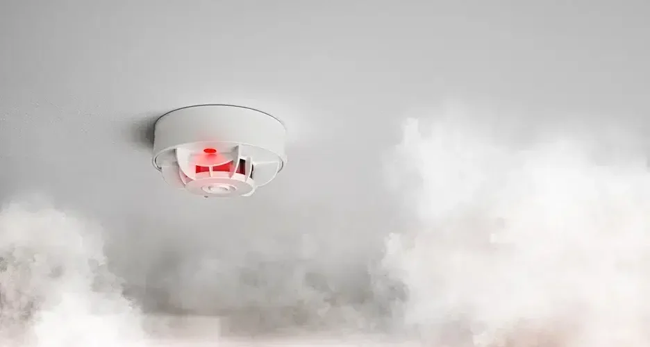Smoke detector activating amid rising smoke on a ceiling.