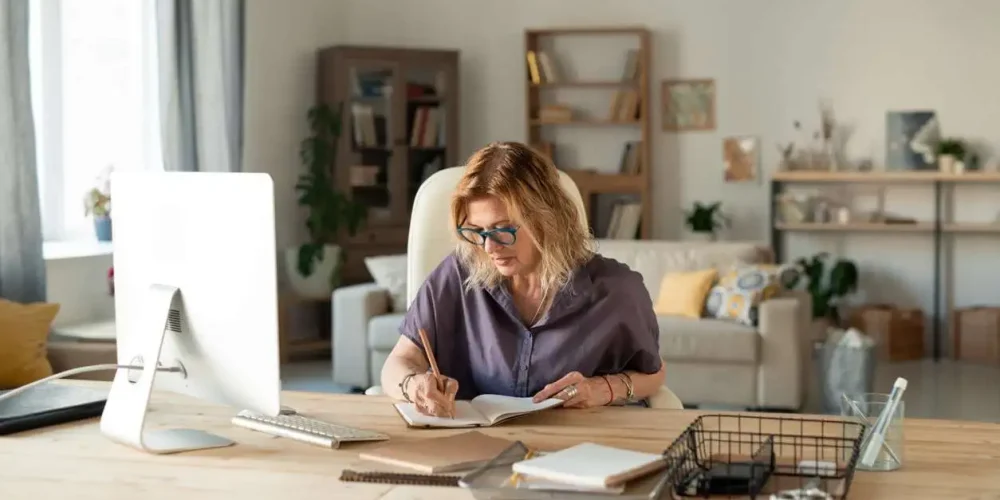 A woman working at a home office desk, writing in a notebook, with a computer and bookshelf in the background.