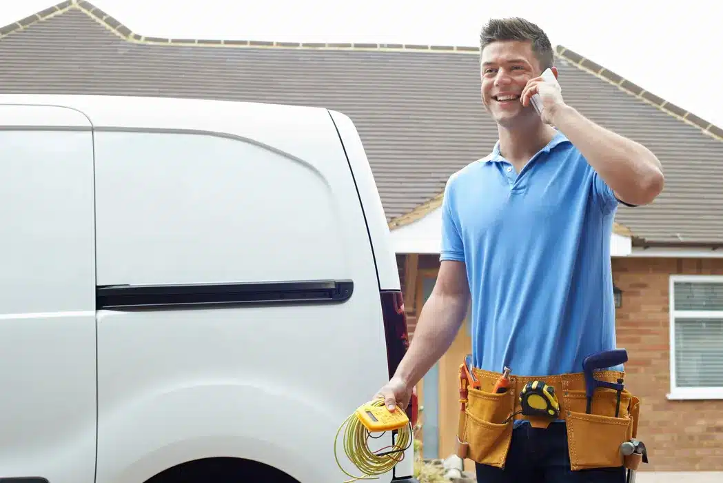 Electrician in blue shirt with tool belt talking on phone beside van outside a house.