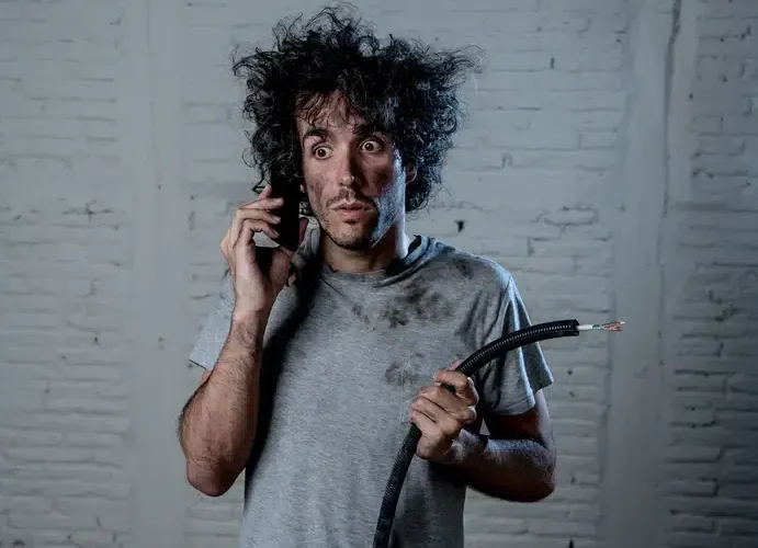A surprised man with messy hair holds a broken cable and phone, wearing a dirty shirt in front of a white brick wall.