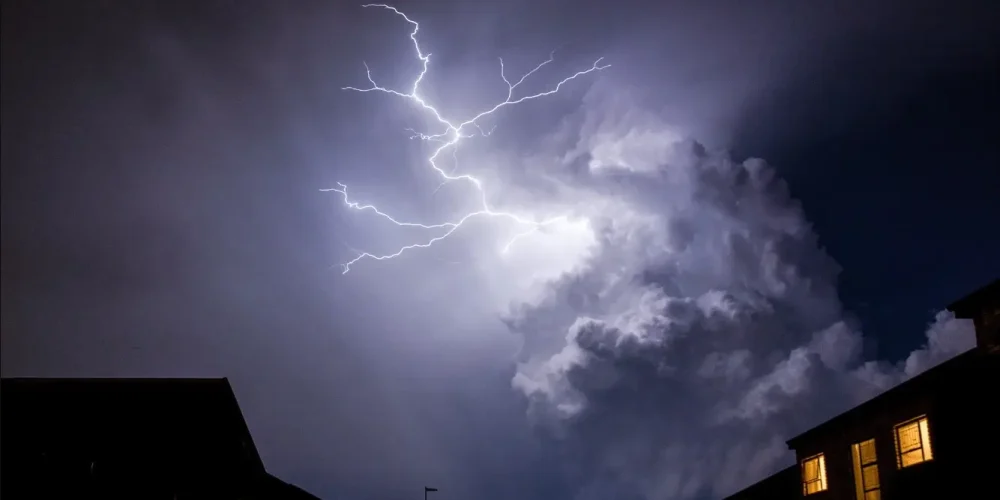 Dramatic lightning bolt illuminates storm clouds over silhouetted buildings at night.