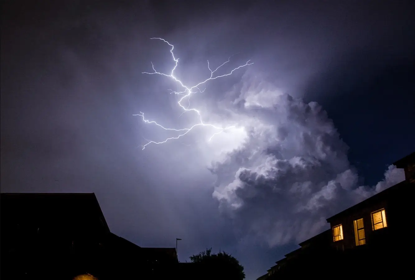 Dramatic lightning bolt illuminates storm clouds over silhouetted buildings at night.