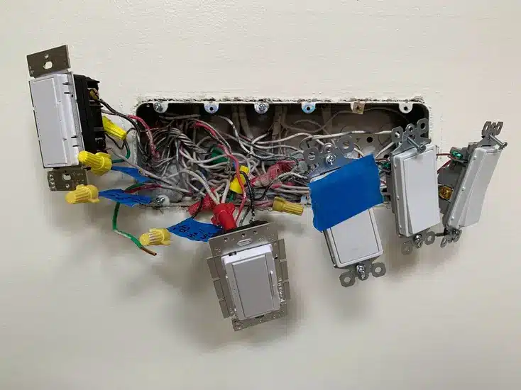 Electrical panel with multiple exposed wiring and light switches on a wall.