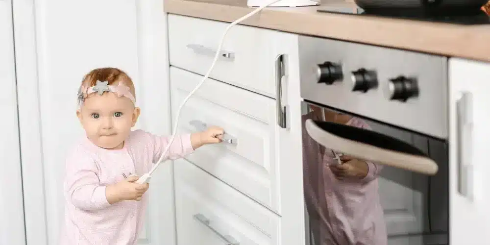 Baby holding a cable, exploring kitchen drawers in a pink onesie, with a decorative headband.