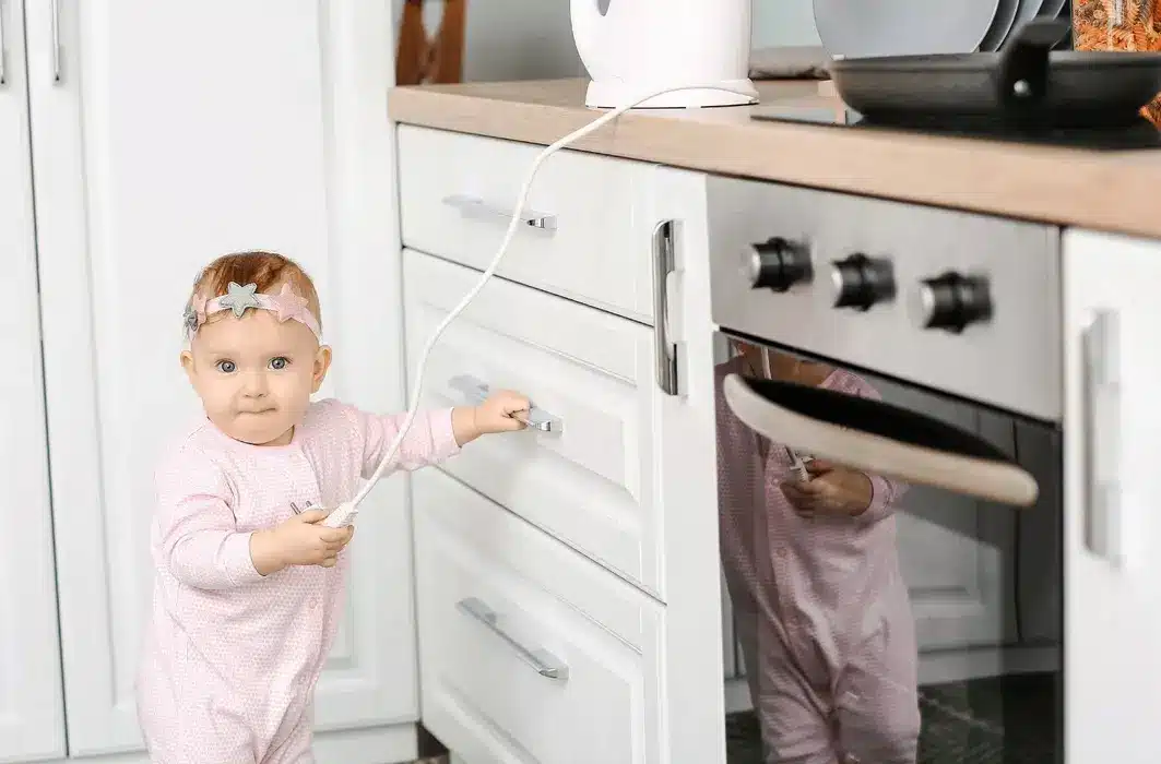 Baby holding a cable, exploring kitchen drawers in a pink onesie, with a decorative headband.