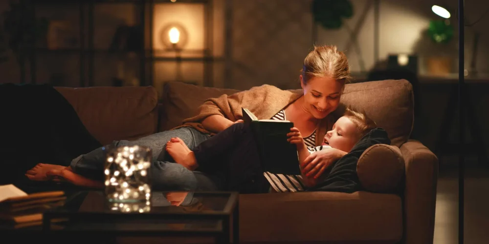 A woman reading a book to a child on a cozy sofa in a warmly lit living room.