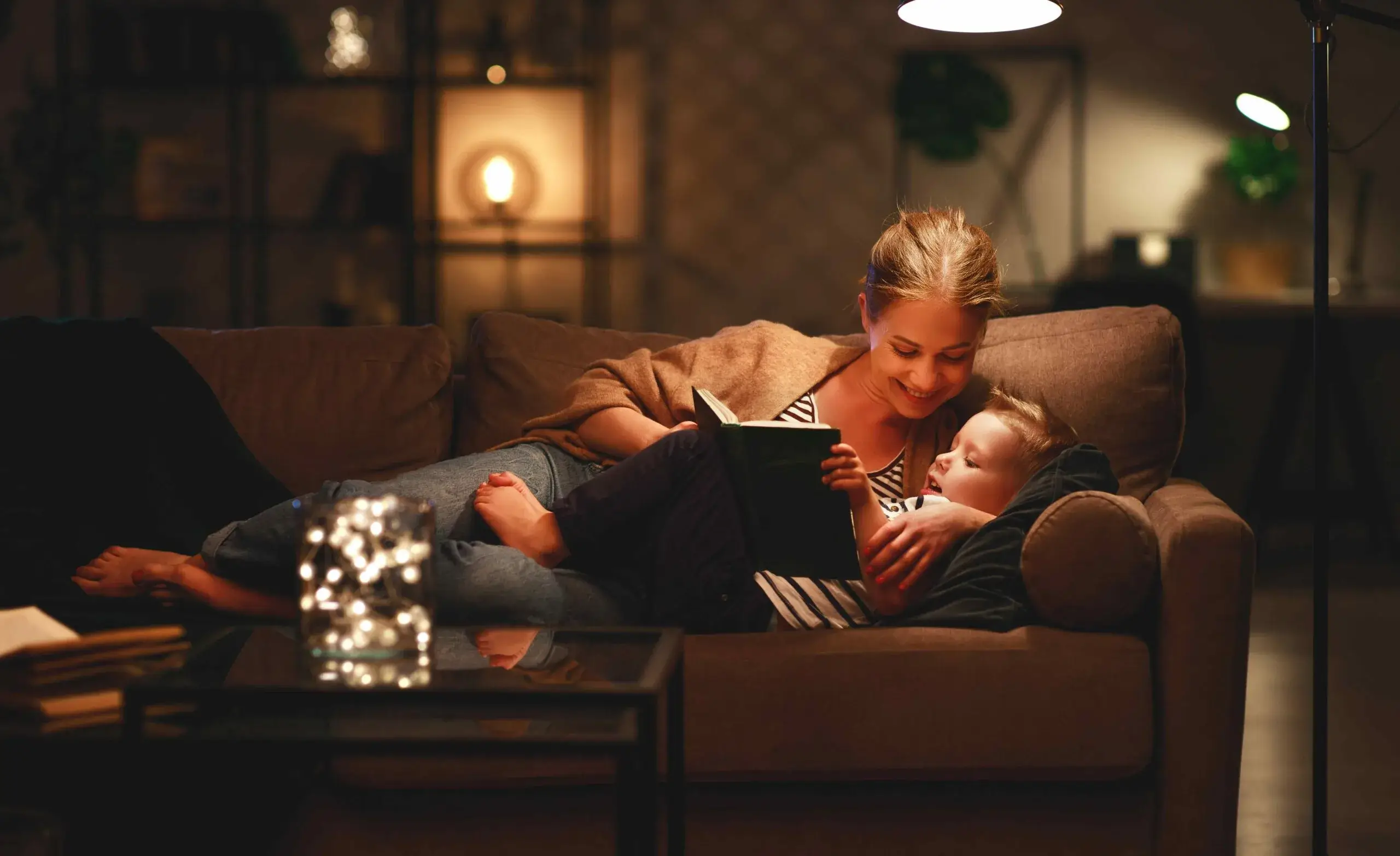 A woman reading a book to a child on a cozy sofa in a warmly lit living room.