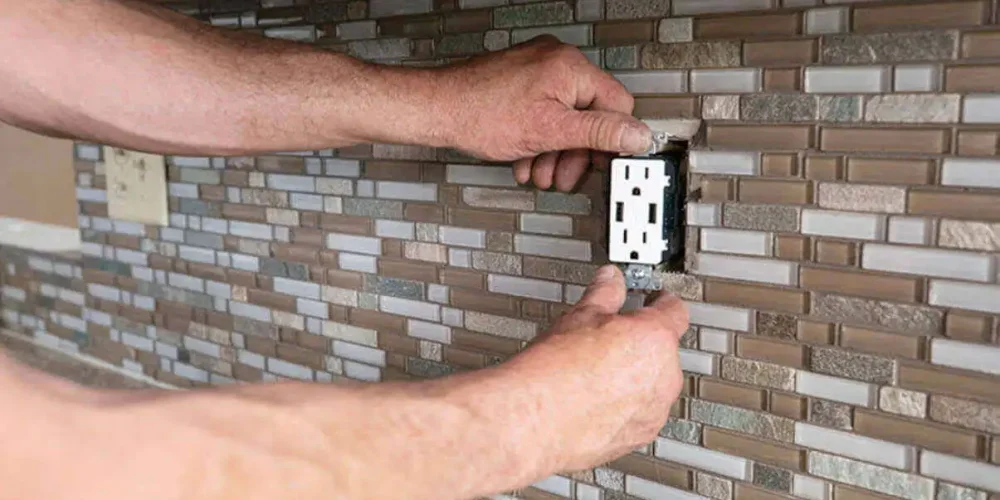 A person installs a power outlet in a tiled kitchen backsplash.