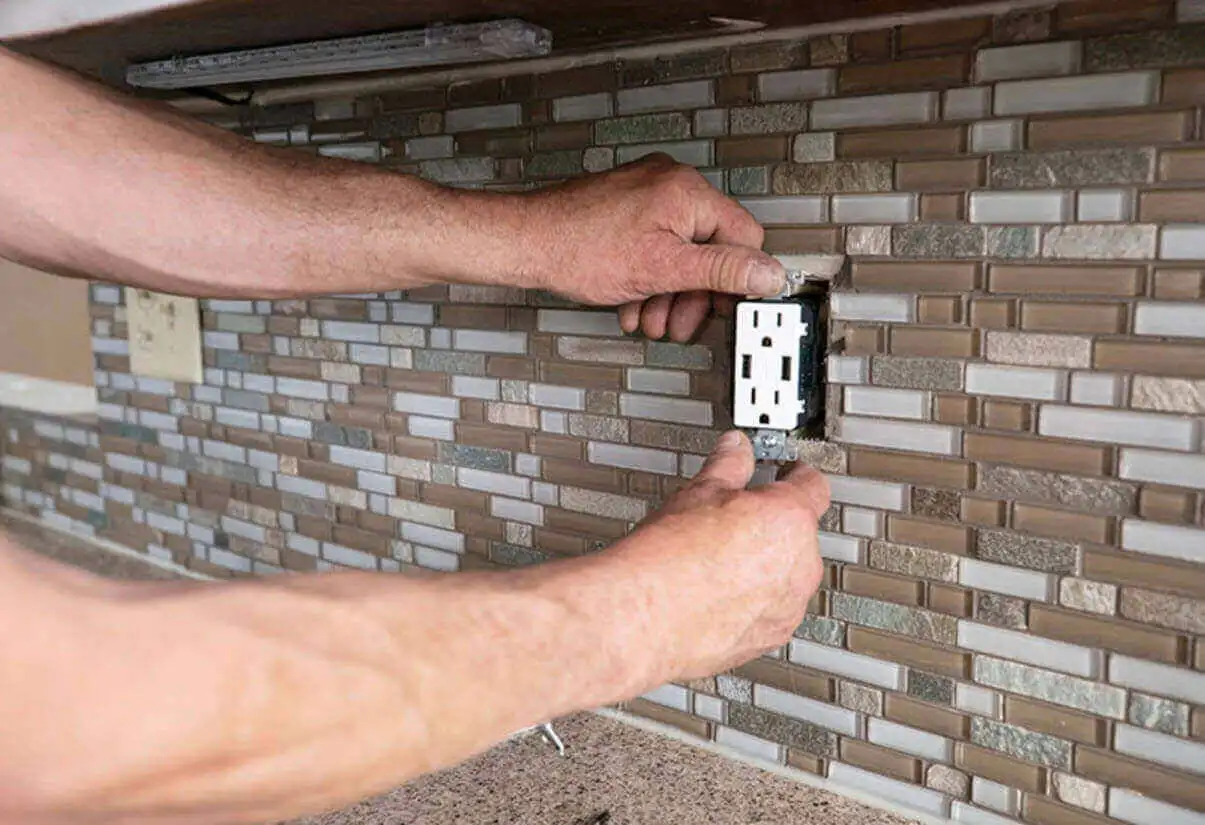 A person installs a power outlet in a tiled kitchen backsplash.