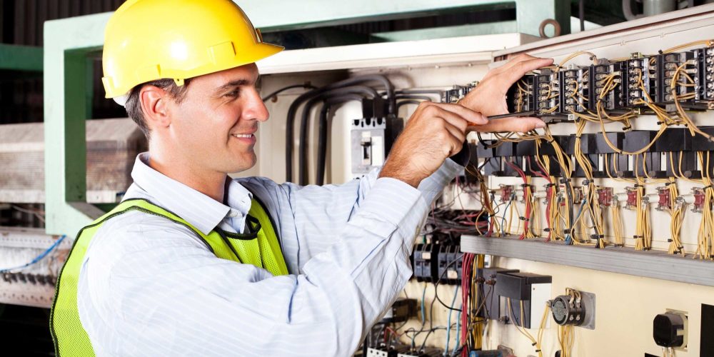 Worker in a hard hat and safety vest, adjusting electrical wiring panel in an industrial setting.