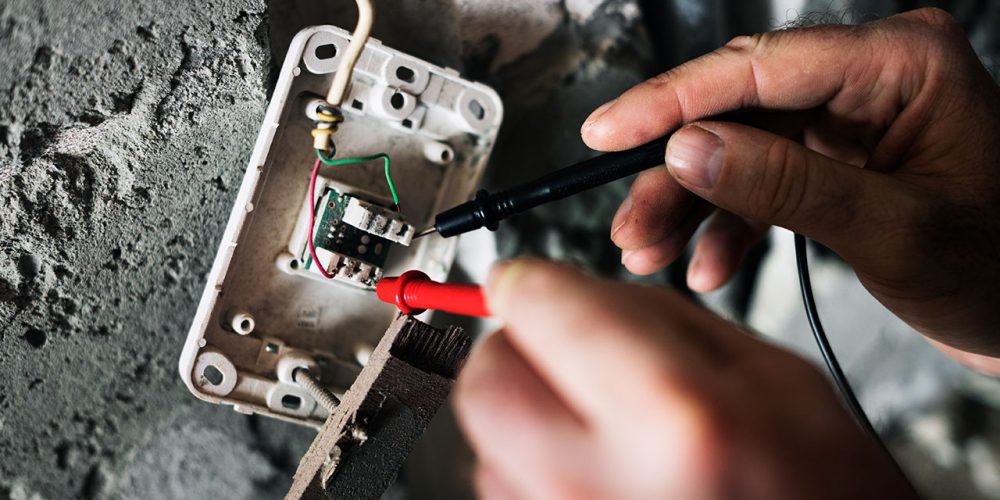 Hands testing electrical wiring with probes in a wall-mounted socket.