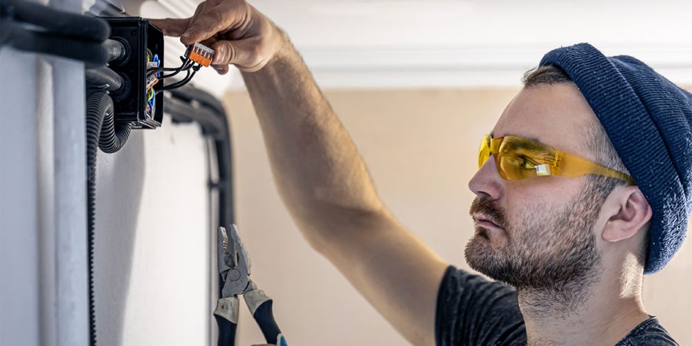 Electrician with safety glasses and beanie fixing wiring on a wall.