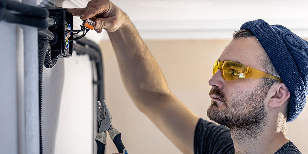 Electrician with safety glasses and beanie fixing wiring on a wall.