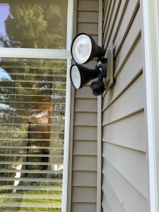 Dual-head motion-sensor security floodlight on house siding beside window with blinds