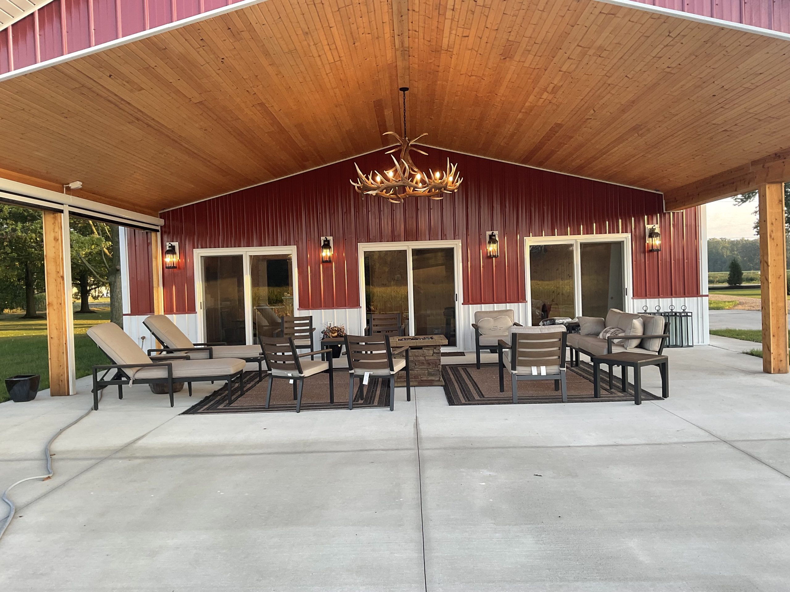 Rustic covered patio at red barn with wood ceiling, antler chandelier, lounge chairs, and fire pit seating